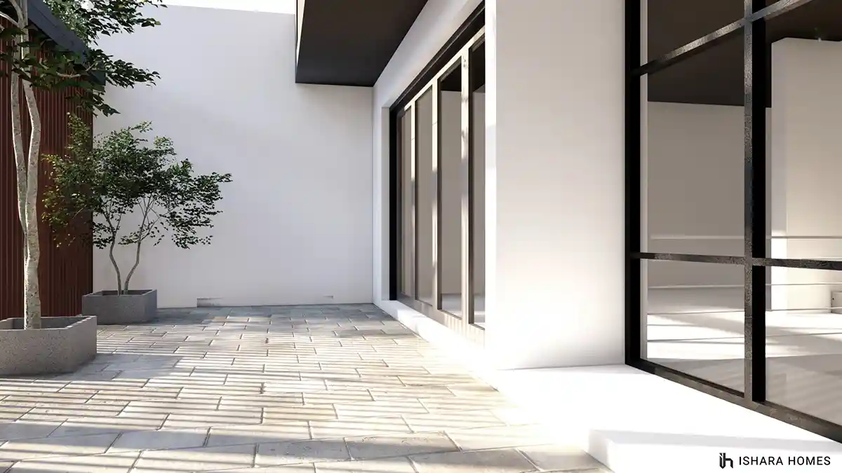 A quiet paved courtyard tucked beside the house, where slim potted trees and a charcoal timber-slat wall meet full-height black-framed glass doors.